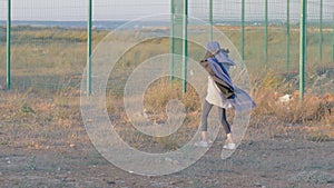 Poor little girl wandering in refugee camp near the state border, strange child walking near the fence covered with blue blanket n