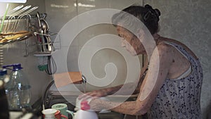 Poor senior woman with sad emotions, gray hair and deep wrinkles, 90 years old, washes and wipes dishes in old kitchen