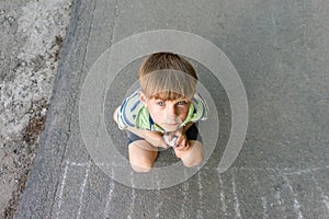 A poor and unhappy boy sits on the asphalt and asks for help while looking into the camera