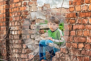 Poor and unhappy orphan boy, sitting on the ruins and ruins of a destroyed building. Staged photo