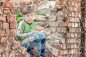 Poor and unhappy orphan boy, sitting on the ruins and ruins of a destroyed building. Staged photo