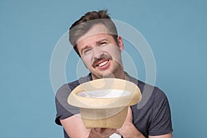 Poor young man holding hat and asking for money over blue background