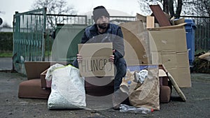 Poorly and dirtyly dressed man sits by a pile of rubbish and holds a poster HUNGRAY in his hands.