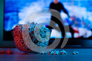 Popcorn in a glass plate on the background of the TV. Color bright lighting, blue and red. Background