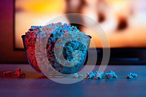 Popcorn in a glass plate on the background of the TV. Color bright lighting, blue and red. Background