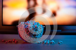 Popcorn in a glass plate on the background of the TV. Color bright lighting, blue and red. Background