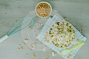 Popcorn in white bowl with pastel spoon, salt, hand mixer and corn seeds on wood background. Cooking for party