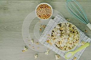Popcorn in white bowl with pastel spoon, salt, hand mixer and corn seeds on wood background. Cooking for summer party