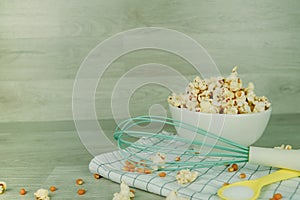 Popcorn in white bowl with pastel spoon, salt, hand mixer and corn seeds on wood background. Cooking for summer party