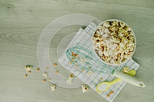 Popcorn in white bowl with pastel spoon, salt, hand mixer and corn seeds on wood background. Cooking for summer party