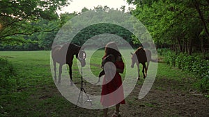 popular blogger is recording videoblog standing in forest meadow with dog and horses on background talking to subscribers.