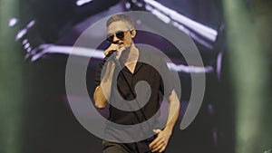 Popular male performer in black round glasses sings into microphone on stage of nightclub in light of bright spotlights