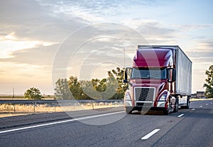 Popular model burgundy big rig semi truck transporting commercial cargo in dry van semi trailer running on the evening highway