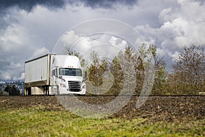 Popular white big rig semi truck transporting cargo in dry van semi trailer running on the road at stormy weather