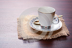 Porcelain coffee cup with saucer and golden spoon on a jute napkin. Vintage coffee set on wooden table