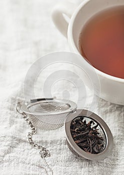 Porcelain cup with hot black tea and infuser with loose tea on white towel
