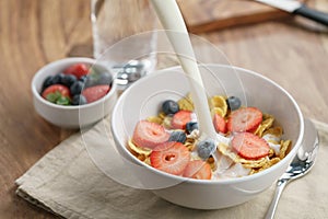 Poring milk into corn flakes with berries in bowl on table