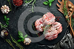 Pork cutlets wrapped in bacon served on a black plate with rosemary and thyme. On a black stone background.