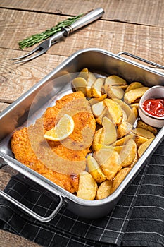 Pork meat weiner schnitzel with potato wedges and tomato sauce in steel tray. Wooden background. Top view