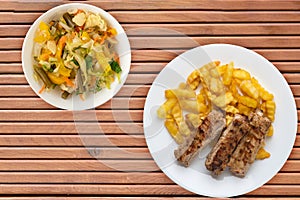 Pork ribs and potatoes on a white plate with salad and sauce on a wooden background. Top view of fast food. unhealthy food