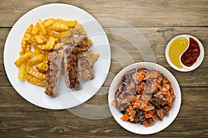 Pork ribs and potatoes on a white plate with salad and sauce on a wooden background. Top view of fast food. unhealthy food