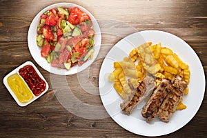 Pork ribs and potatoes on a white plate with salad and sauce on a wooden background. Top view of fast food. unhealthy food