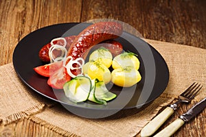 Pork sausages with boiled potatoes, cucumber, and tomato, served on a black plate.