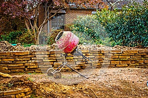 Portable cement mixer at landscaping site by rock wall being built near brick house with Japanese Maple tree and lush vegetation