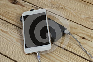 Portable charger charges a smartphone on a wooden table. Mobile phone mockup with dark screen and power bank