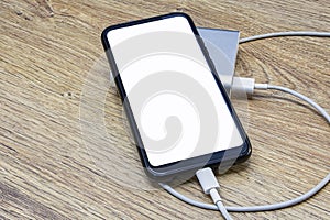 Portable charger charges a smartphone on a wooden table. Mobile phone mockup with white screen and power bank