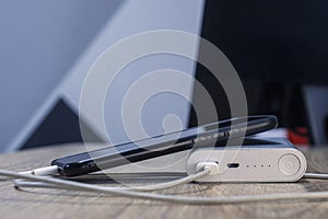 A portable charger charges a smartphone on a wooden table. Power bank with phone and cable on the background of the monitor