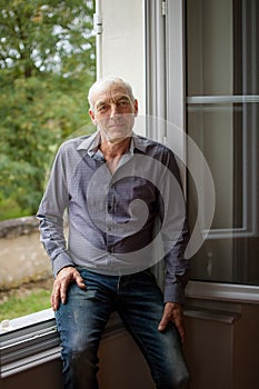 Portait of Handsome Senior Grey-Haired Man Looking at the Camera Standing in Front of Opened Window in His House.