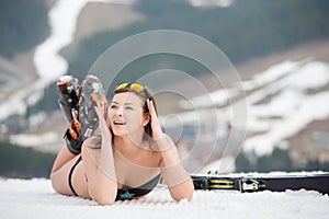 Portait of woman skier is relaxing on the snowy slope in the mountains. Wearing swimsuit, boots and sunglasses