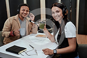 Portait of two cheerful radio hosts, man and woman smiling at camera, drinking coffee or tea while getting ready for a