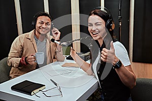 Portait of two cheerful radio hosts, man and woman smiling at camera, drinking coffee or tea while getting ready for a