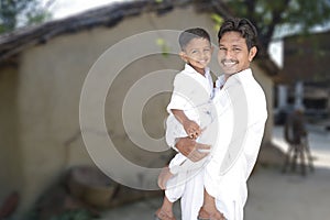 Portarait of happy smiling rural indian young father and little cute son in white traditional cloths with village background, copy