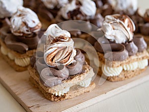 Portion cakes of meringue and cocoa butter cream in square form on a wooden stand, a piece of cake on a light wooden background.