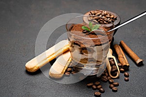 Portion of Classic tiramisu dessert in a glass cup, scoop full of coffee and cinnamon sticks on dark concrete background