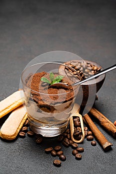 Portion of Classic tiramisu dessert in a glass cup, scoop full of coffee and cinnamon sticks on dark concrete background