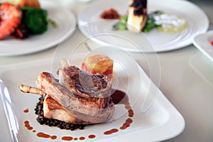 Portion of grilled lamb chop rib on white square plate at restaurant other foods at background