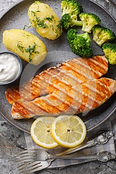 Portion of grilled salmon with boiled potatoes, broccoli, lemon, herbs and cream sauce close-up in a plate on the table. Vertical