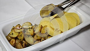 Portion potatoes of two types on one plate. Rectangular white plate with iron tongs for serving rustic potatoes with