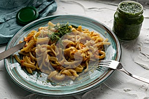 Portion of spaghetti pasta like noodles with fried vegetables on a modern plate in turquoise color, with green sauce in a jar, the