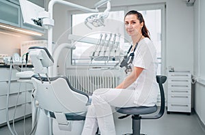 Portraait of sincerely smiling young dentist woman dressed white medical scrubs uniform sitting in modern dental clinic next to