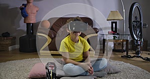 Portrair of laughing Caucasian boy in VR googles sitting on the carpet and moving his body. Happy schoolboy in using