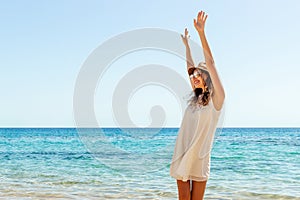 Portrair of young woman dressed in a white dress and a brown hat standing at the beach and putting hads up. Girl is engoying her