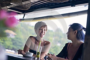 Portrait of 2 asian women chatting, drinking & smiling at beach bar in summer