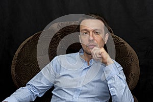 Portrait of a 30-35-year-old man in a blue shirt, sitting in an armchair on a black background. Perhaps he is a lawyer or a lawyer