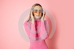 Portrait of 30-35 years woman in bright pink dress touching sunglasses with hands on pink background. Studio shot