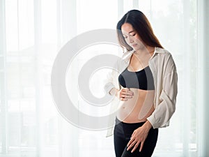 Portrait of 4 months pregnant Asian woman in white bed room, Woman touching her abdomen belly on white background.